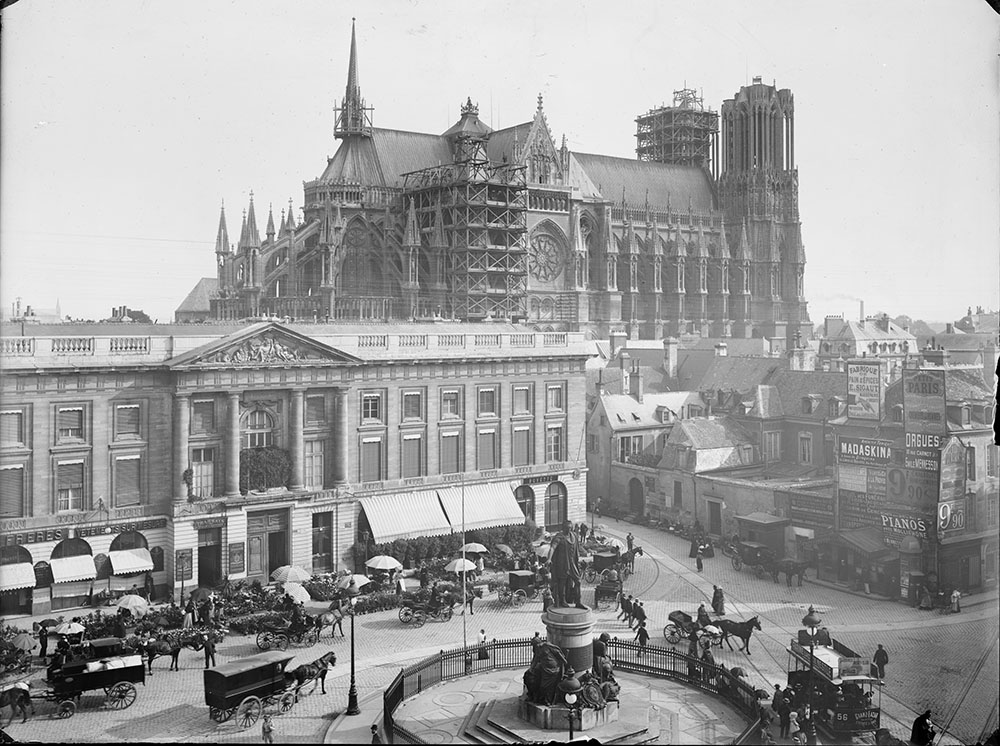 Vue plongeante sur la place Royale avec la cathédrale en arrière-plan. La présence du tramway hippomobile, qui côtoie les calèches à chevaux, ainsi que les divers commerces qui longent la place nous permettent de plonger dans Reims telle qu’elle était il y a plus d’un siècle. - BM Reims, PDV Sainsaulieu 64